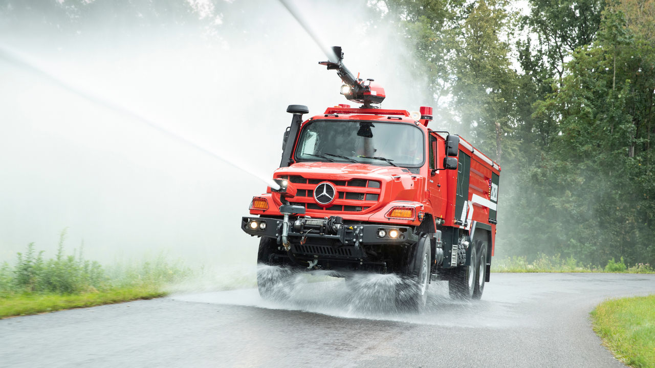 A Unimog airport fire-fighting vehicle drives on a road, using two water cannons to dispense large quantities of water or extinguishing foam. The first cannon is located on the vehicle roof, the second at the height of the headlights. The spray indicates a high speed.