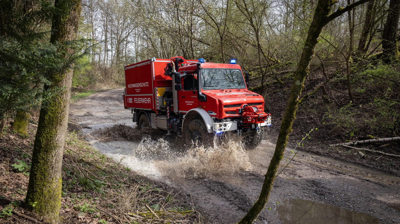  A red fire service Unimog drives on a muddy forest road. The flying spray indicates the high speed and off-road capability of the vehicle. The body of the vehicle is designed for forest fire fighting.