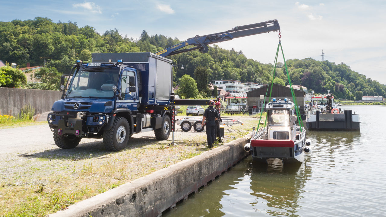A dark blue Unimog with a crane body lifts a vessel out of the water. Two people in uniform are standing on the shore, securing the manoeuvre. A river and wooded hills can be seen in the background.