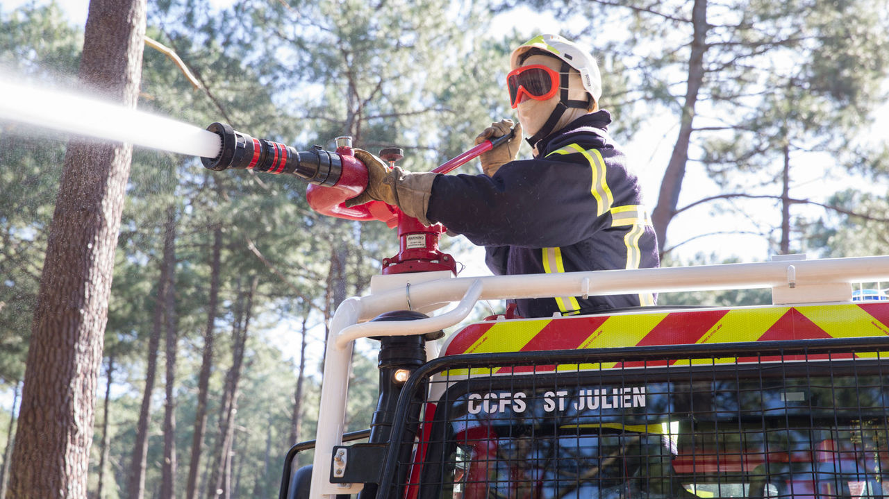 A firefighter in protective clothing including helmet, goggles and face shield operates a water canon on a fire-fighting vehicle. The jet of water shoots into the left half of the image; trees are visible in the background. The vehicle has “CCFS ST JULIEN” written on it.