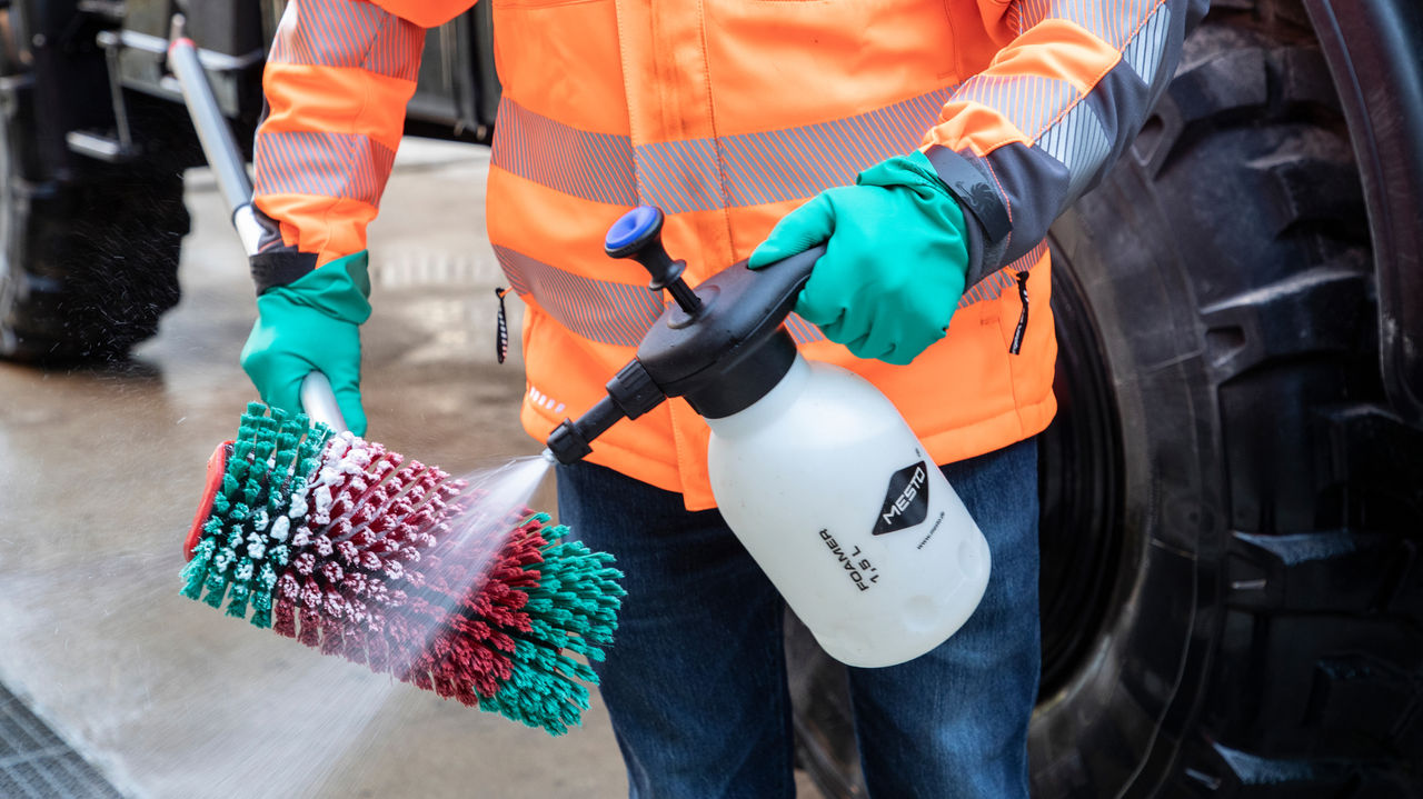 Une personne portant une veste de travail orange pulvérise du produit de nettoyage sur une brosse verte et rouge fixée à un manche télescopique.