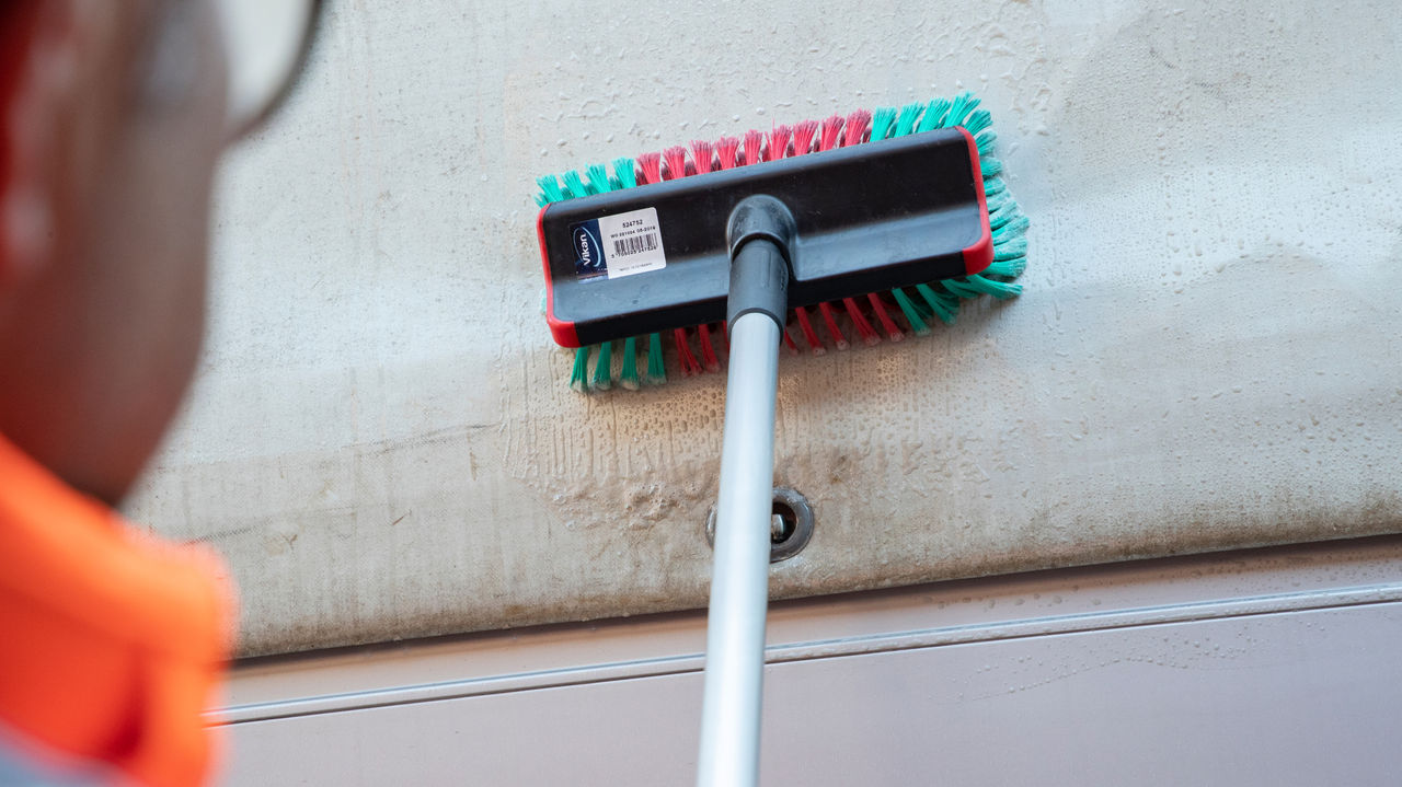 Une personne en veste de travail orange frotte la surface extérieure d'une bâche de camion avec une brosse montée sur un manche télescopique.