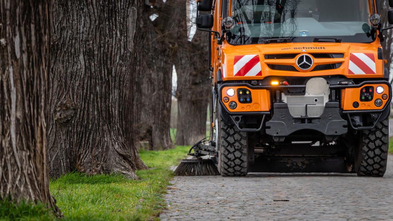 The image shows a close-up of an orange Unimog with a road sweeper on a cobbled road. The rotating brush on the side of the machine is in operation, sweeping the road. Large tree trunks and a green strip of lawn can be seen in the foreground.