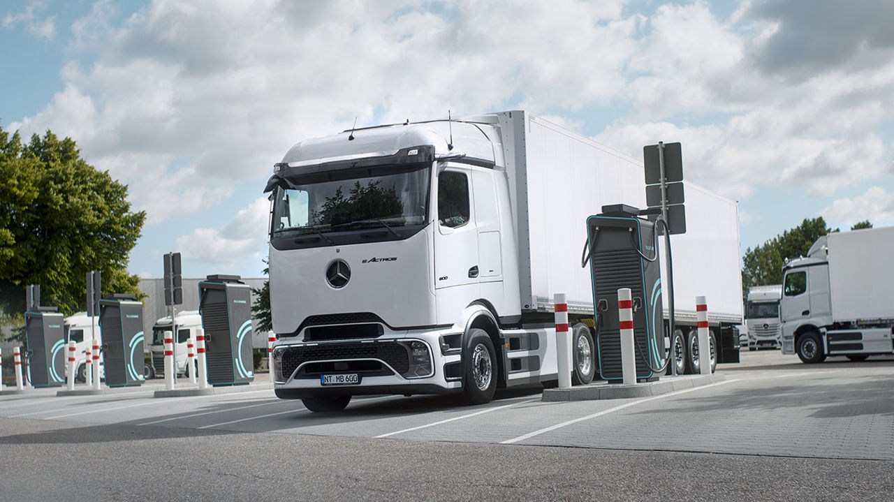 Un eActros 600 blanc avec ProCabin de Mercedes-Benz Trucks, accompagné d'une remorque blanche, est stationné sur un parking et en cours de recharge à une borne de recharge. Plusieurs bornes de recharge et poteaux de délimitation sont visibles autour du camion. En arrière-plan, d'autres camions blancs et, sur le bord gauche de l'image, des arbres verts sous un ciel partiellement nuageux.