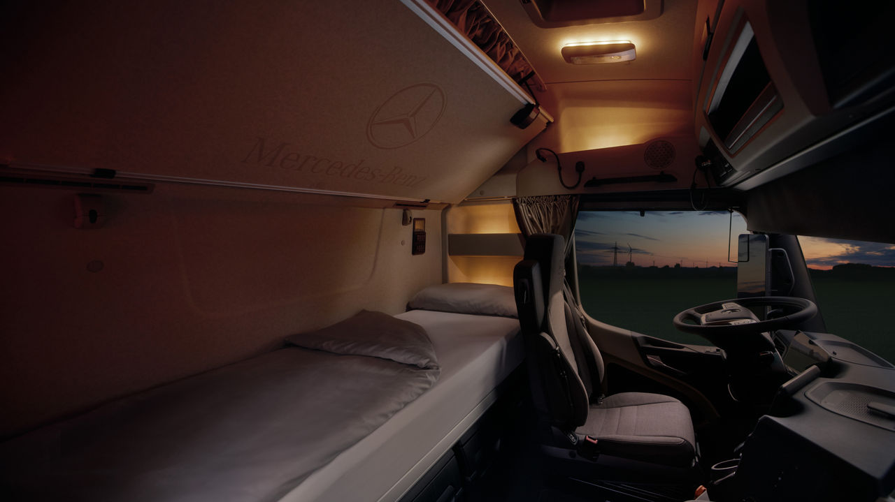L'image montre l'intérieur de la cabine d'un eActros de Mercedes-Benz Trucks au crépuscule. À gauche, on peut observer un lit soigneusement fait avec des draps gris clair, et au-dessus, au plafond, le logo Mercedes-Benz et l'inscription « Mercedes-Benz ». À droite se trouvent le siège conducteur et le volant, tandis que le pare-brise laisse entrevoir un ciel sombre avec les silhouettes d'éoliennes et un vaste paysage.