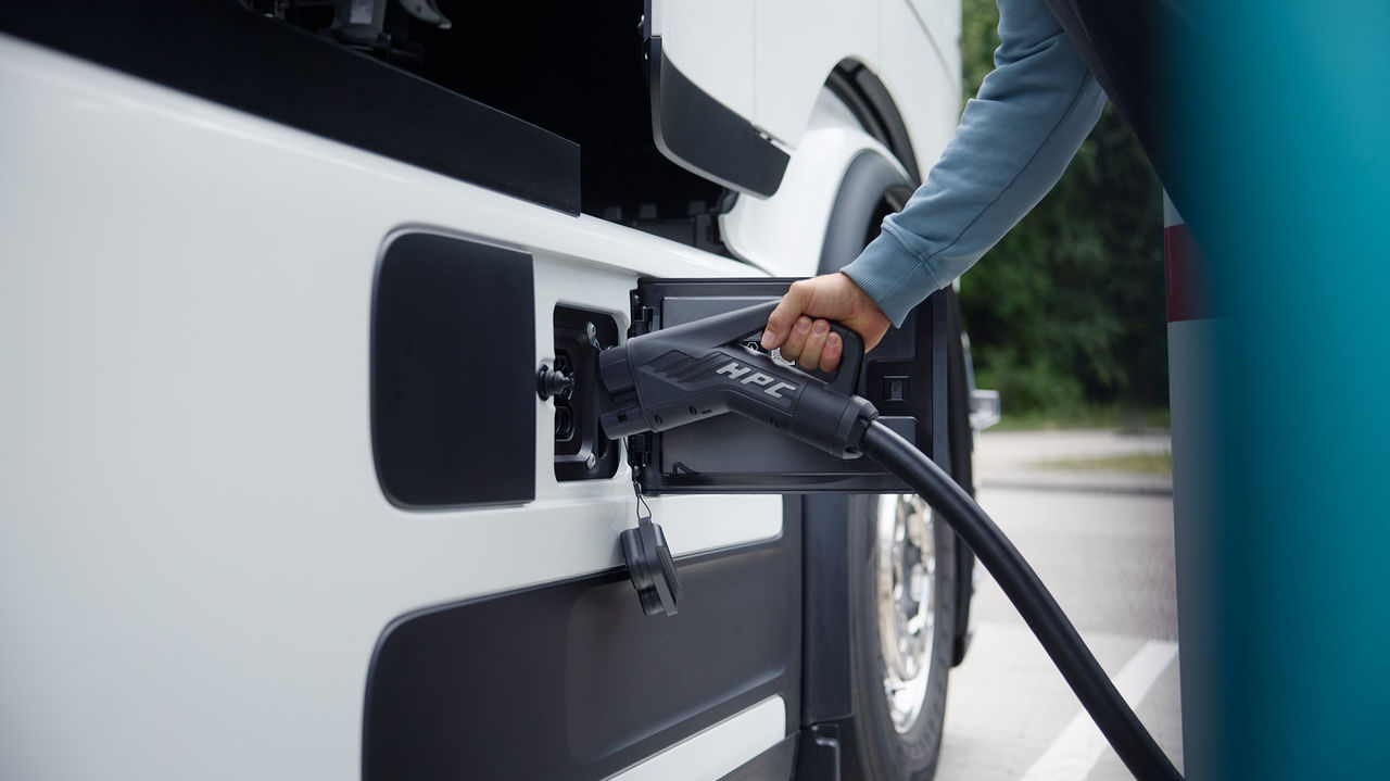 Une personne branche un câble de charge dans l'eActros de Mercedes-Benz Trucks, qui est blanc et partiellement visible sur l'image. L'accent est mis sur le processus de charge, le câble et le connecteur étant clairement visibles.