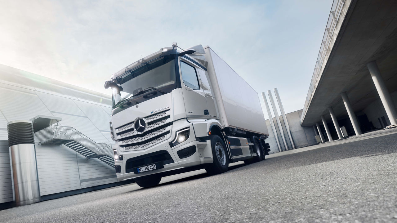 Un eActros 400 blanc de Mercedes-Benz Trucks est stationné sur une surface asphaltée devant un bâtiment moderne doté d'un escalier extérieur, avec un éclairage solaire intense provenant du haut à gauche. À droite de l'image, on peut observer une structure en béton surélevée avec des colonnes, qui suggère un passage souterrain ou une zone couverte.