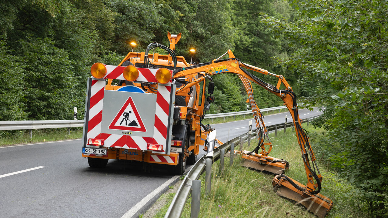 An orange Unimog with an extendable mower arm mows by the roadside. The vehicle has a red and white warning sign and a construction site sign at the rear. The mower arm is leaning over the crash barrier and mowing the vegetated side of the slope next to the road. A forested landscape can be seen in the background.