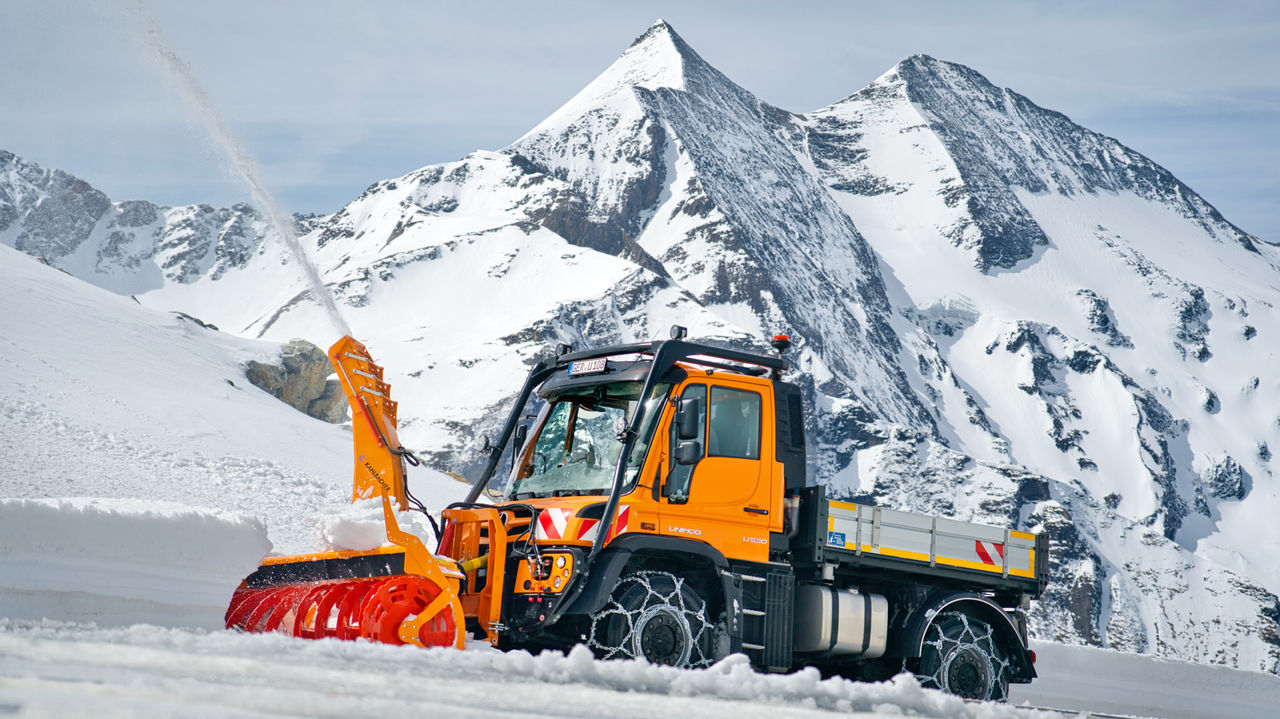 The image shows an orange Unimog with a snow blower clearing a snow-covered track in the mountains. The snow blower throws the snow high into the air. Snow chains are fitted on the tyres. Snow-covered mountain peaks are visible in the background.