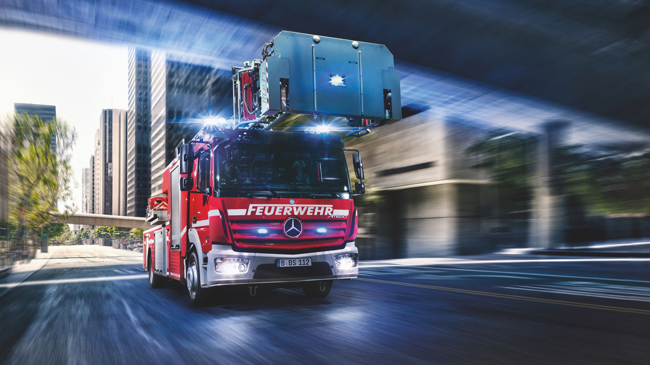 Un camion de pompiers Atego rouge roule rapidement dans une ville avec les feux bleus allumés. L'échelle du véhicule est déployée. En arrière-plan, on voit des gratte-ciel et un pont. L'image donne un sentiment d'urgence et de mouvement.