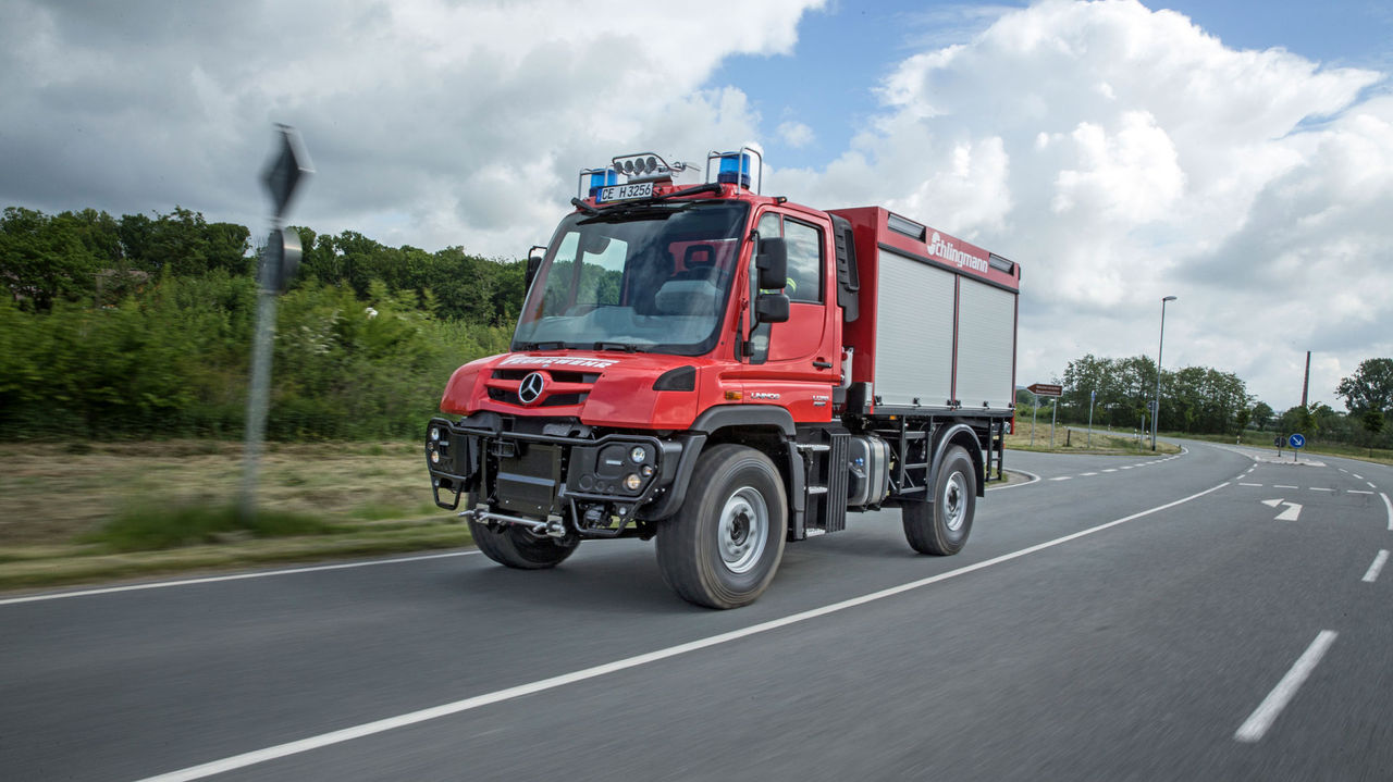 Un Unimog roule sur une route avec un gyrophare. Le véhicule est peint en rouge et possède une superstructure de pompiers. En arrière-plan, on aperçoit des arbres et des buissons. 