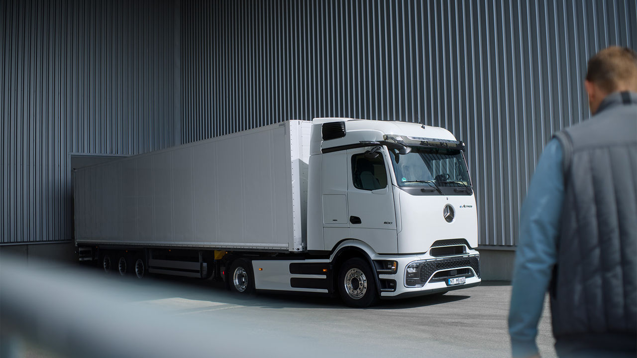 Un eActros 600 blanc avec ProCabin de Mercedes-Benz Trucks, accompagné d'une longue remorque blanche, est stationné sur un terrain asphalté devant un mur de tôle ondulée grise. Le camion est vu de côté, avec l'avant légèrement orienté vers la gauche. Au premier plan, dos à l'observateur, une personne en veste bleue et gilet gris regarde le camion. La lumière éclaire la scène par le haut.