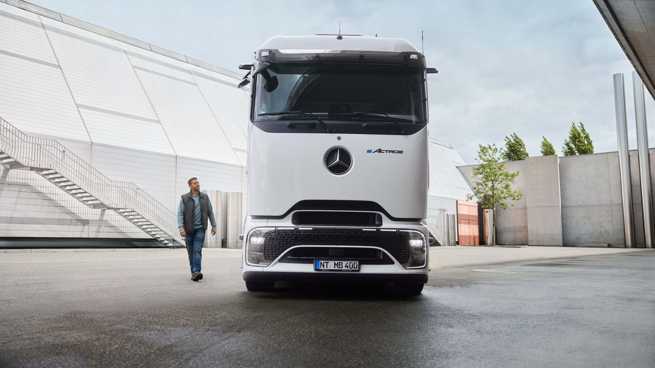 Un homme en tenue de travail marche sur une surface asphaltée, tandis qu'un eActros 400 blanc avec ProCabin de Mercedes-Benz Trucks est garé à l'arrière-plan. À gauche, on peut voir un bâtiment moderne avec un long escalier extérieur et à droite, des arbres et d'autres structures de bâtiment.