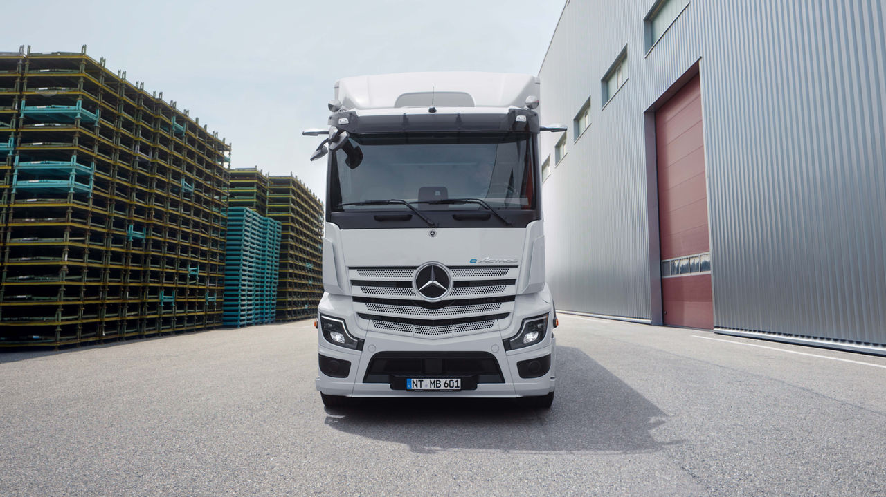 Un eActros 600 de Mercedes-Benz Trucks est stationné face à un bâtiment moderne doté d'un long escalier extérieur, sur une surface asphaltée. Le camion est blanc et arbore l'étoile Mercedes sur la calandre, tandis que des arbres et d'autres structures de bâtiments sont visibles en arrière-plan.