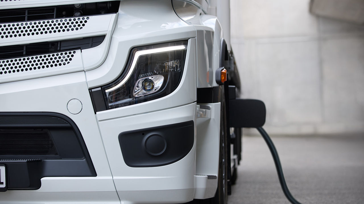 Un eActros 600 de Mercedes-Benz Trucks est photographié depuis le côté droit, l'accent étant mis sur la partie avant du camion. Le véhicule est de couleur blanche et un câble de recharge est branché sur le côté du camion, ce qui indique que la batterie est en cours de recharge.
