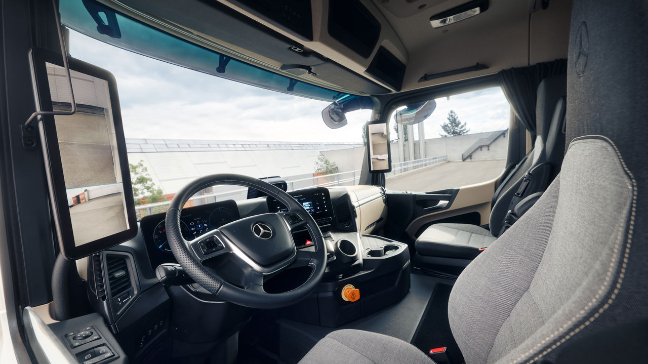 L'image montre l'intérieur de l'eActros de Mercedes-Benz Trucks vu depuis le siège passager. On peut voir le volant avec l'étoile Mercedes, la planche de bord avec les affichages et les écrans numériques ainsi que les sièges confortables en tissu gris clair.