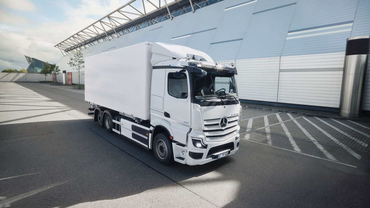 Un eActros 400 blanc de Mercedes-Benz Trucks avec une caisse est stationné sur un parking asphalté devant un bâtiment moderne doté d'une façade en verre. La lumière du soleil projette de longues ombres sur le sol, ce qui indique une journée claire.