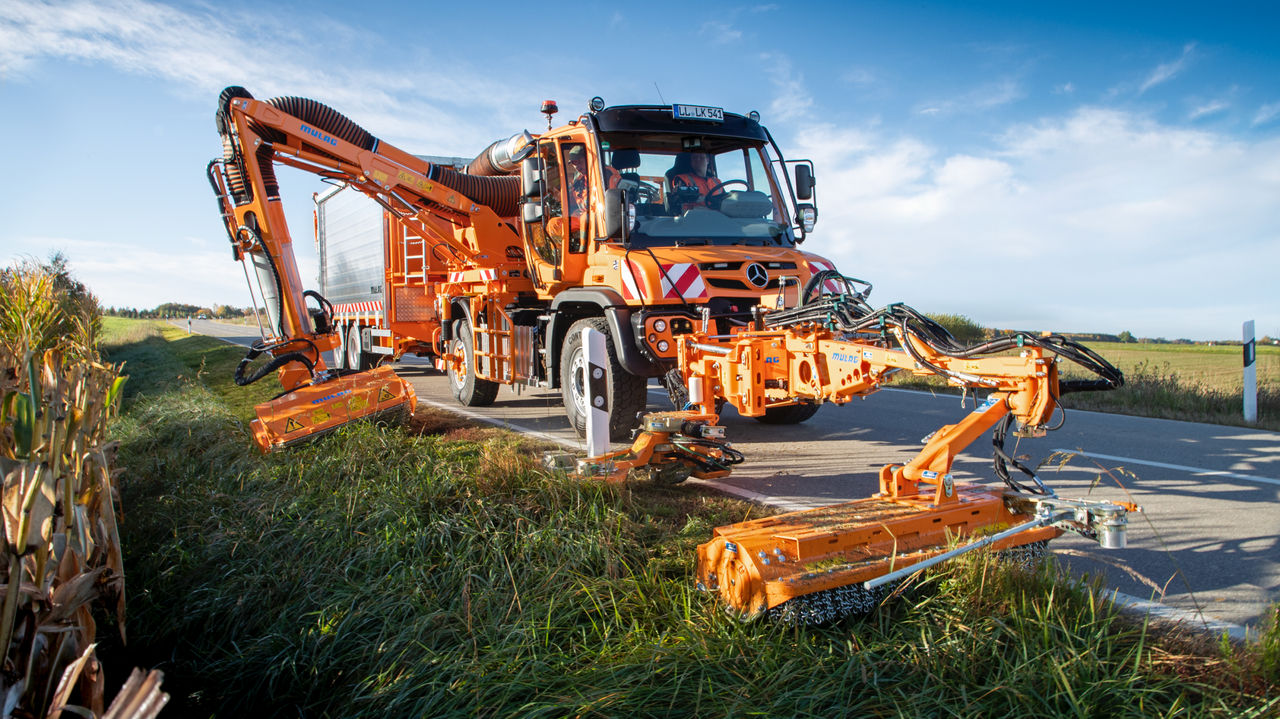 An orange Unimog with two side-extended mower decks works on the roadside. The Unimog, with a boom-like body behind the cab, is driving on an asphalt road. Tall grass and corn plants can be seen next to the road. 