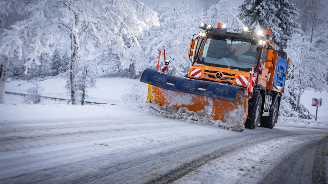 An orange Unimog snowplough drives on a snow-covered road, clearing snow with its blue-orange snow blade. The road is covered with a mixture of snow and mud, and has lane markings. Snow-covered trees line the edge of the road. 