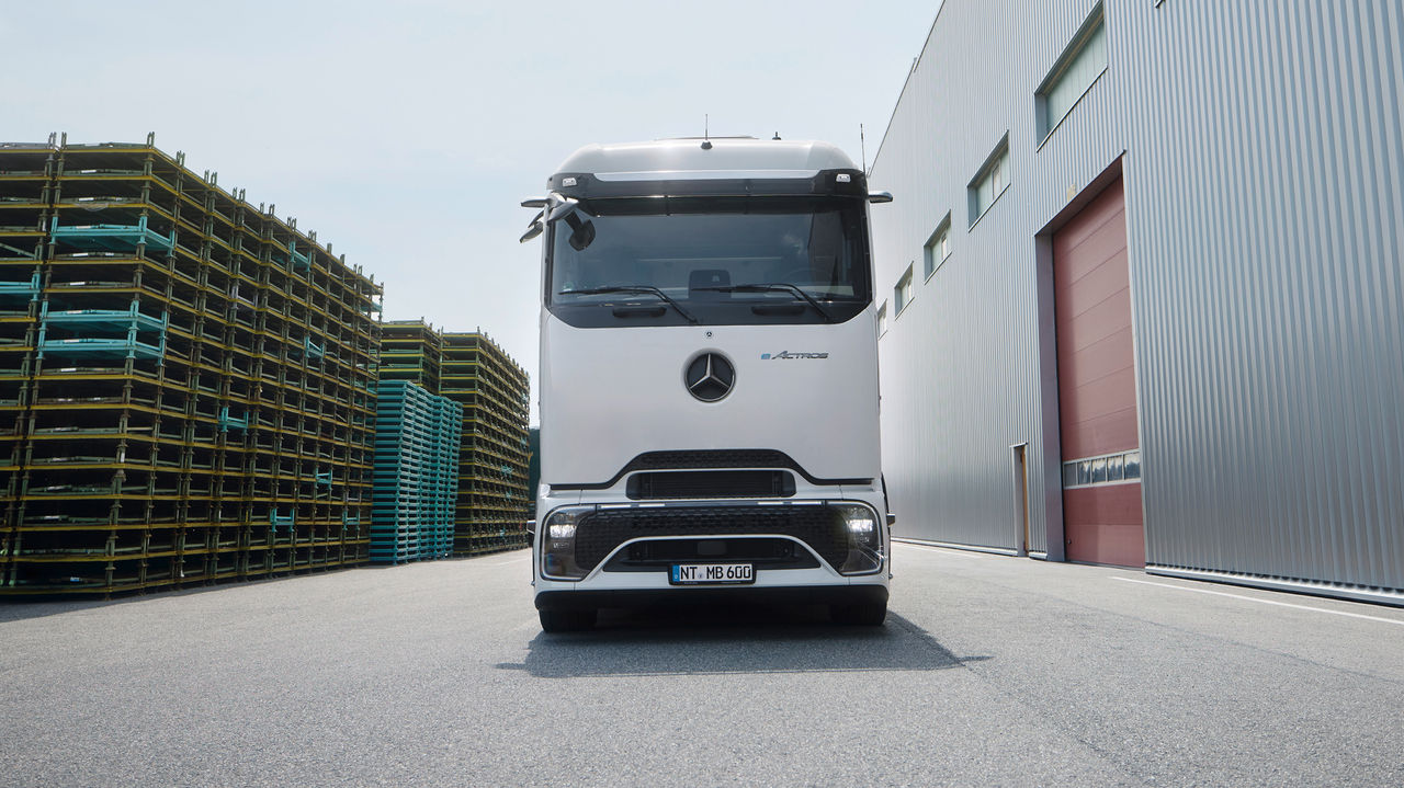 An eActros 600 with ProCabin from Mercedes‑Benz Trucks is facing the camera on an asphalt‑paved area. On the left of the image are tall stacks of yellow and blue pallets, while on the right stands a large, modern building with a grey corrugated‑metal facade and a red gate. The sky is bright and slightly cloudy.