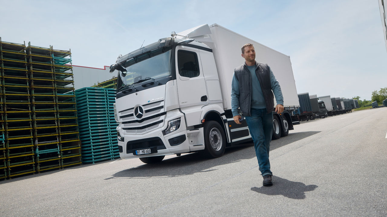 A man in work clothes walks on a paved surface, while in the background a white eActros 600 from Mercedes‑Benz Trucks with a trailer is parked. Stacked pallets are visible on the left, and on the right a large building suggests a warehouse or industrial complex.