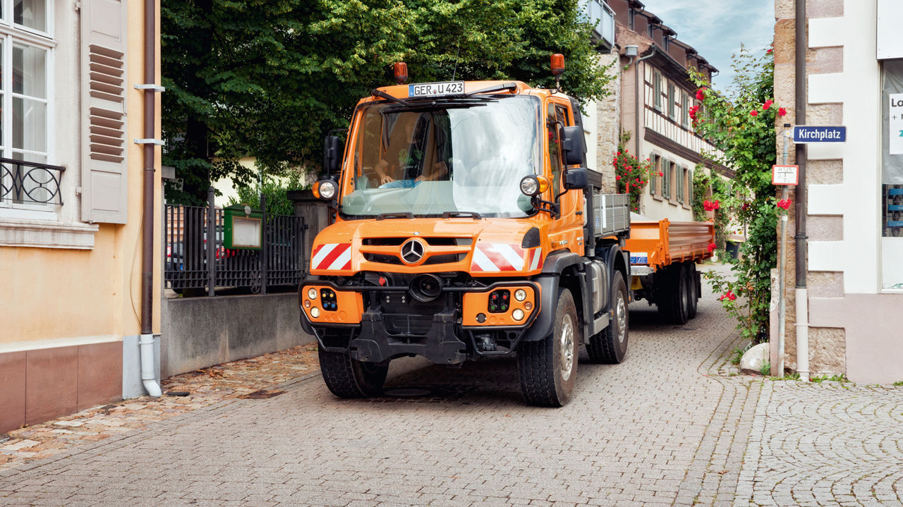شاحنة Unimog برتقالية مع مقطورة تسير في شارع مرصوف داخل إحدى المدن.