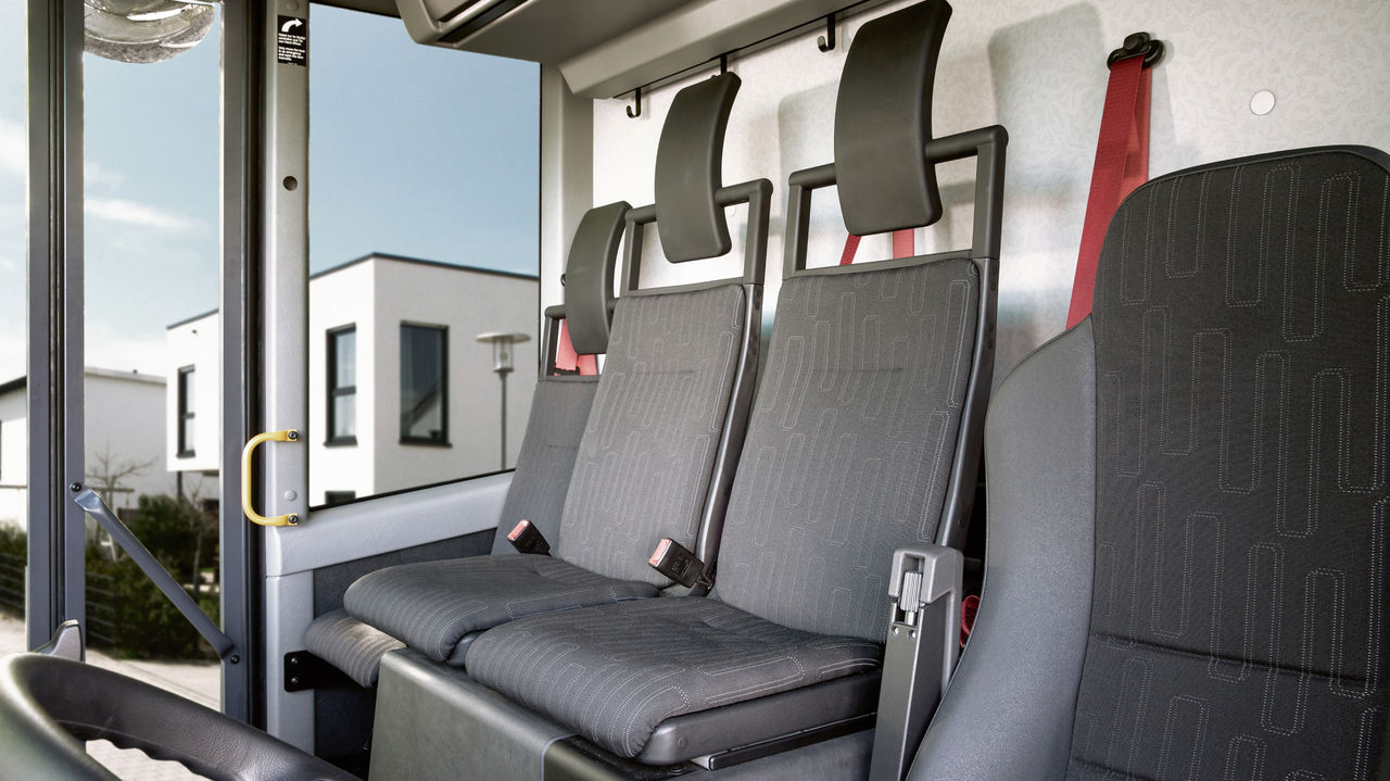 Close-up of three empty passenger seats. The seats are in grey fabric with black head restraints and red seat belts.