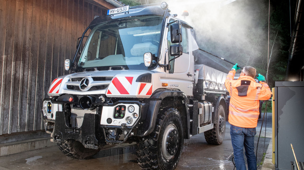 رجل يرتدي سترة عمل برتقالية ينظف شاحنة Unimog فضية باستخدام منظف ضغط عالي. وهو يقوم برش نافذة السائق. يمكن رؤية الرذاذ فوق الشاحنة.