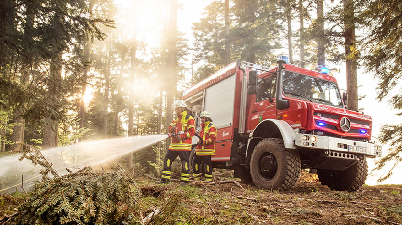 The image shows a red Unimog fire-fighting tanker in operation in the forest. Two firefighters wearing protective clothing use a hose to extinguish a small fire or embers on the ground. The Unimog stands on a forest path surrounded by trees in the sun.