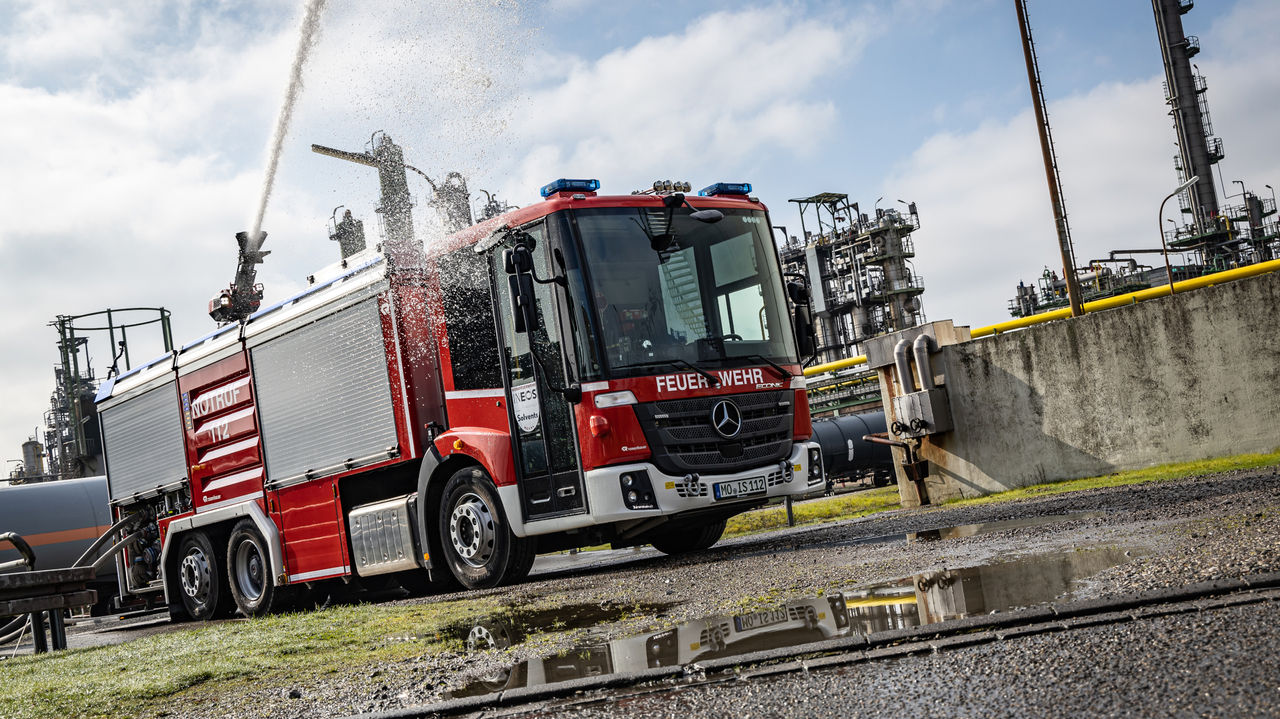 A red Econic fire truck is in operation on the premises of an industrial plant. The vehicle has an extendable water cannon on the roof firing a jet of water high into the air. The industrial plants can be seen in the background.