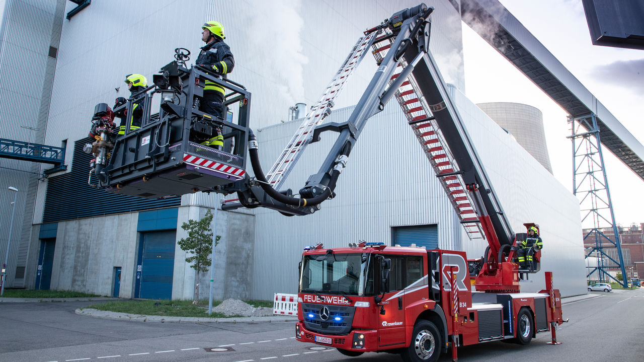 A red Econic with an extended articulated mast stands in front of an industrial building. There are two firefighters in the cage of the aerial rescue platform, which is being operated by another crew member. The scene takes place in front of an industrial building.