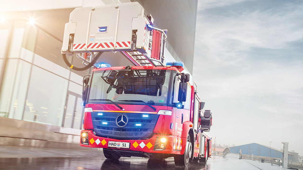 A red Econic with an aerial rescue platform drives to an incident on a rainy road, surrounded by industrial buildings. The blue lights of the fire-fighting vehicle are switched on.