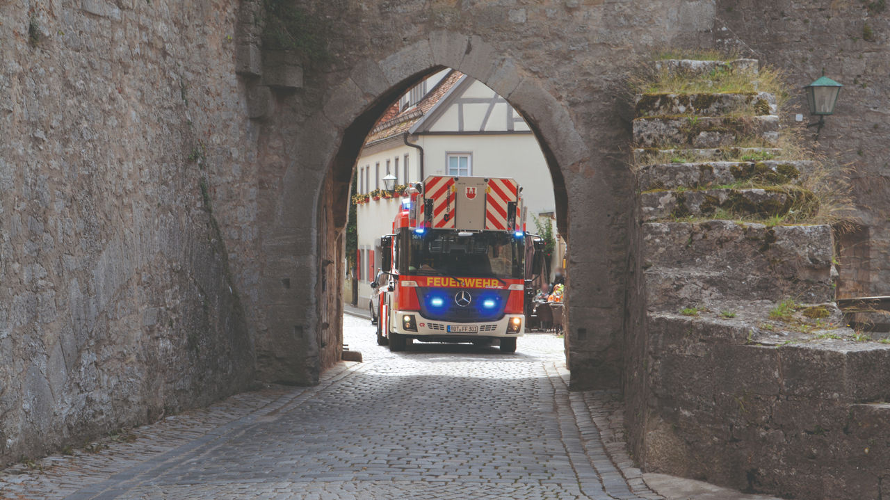 A red Econic with its blue lights on drives through an old city gate on a cobbled road. The aerial rescue platform is retracted. The blue lights of the Unimog are switched on and historic buildings can be seen in the background.