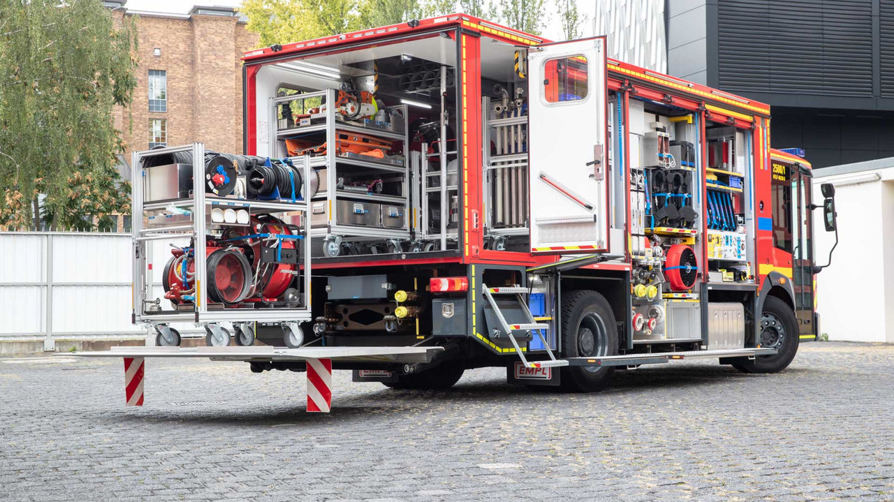 A red fire service Econic with open roller shutters stands on a cobbled square. The body of the equipment truck shows a variety of equipment items and devices that are accessible via an extendable loading surface.