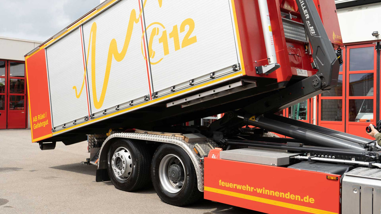 The image shows a red truck in front of the fire service garage. A tipper system vehicle with a fire-fighting container raises its loading surface. It has "feuerwehr-winnenenden.de" written on it in yellow.