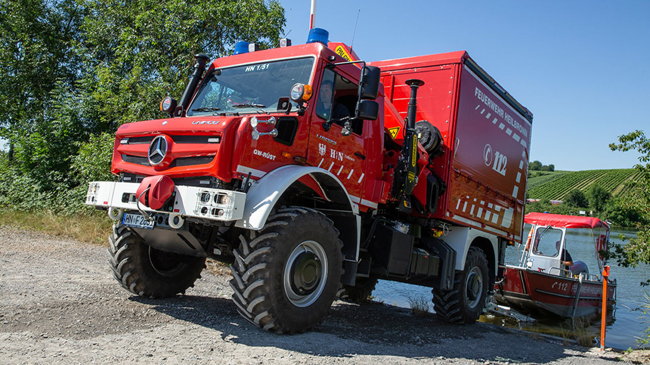 A red Unimog stands on the shore of a body of water and has launched a small, red fire-fighting vessel. The Unimog has a traditional fire service body and seems to be equipped for water rescue operations. It is standing on a gravel path. In the background, you can see vines, water and trees.