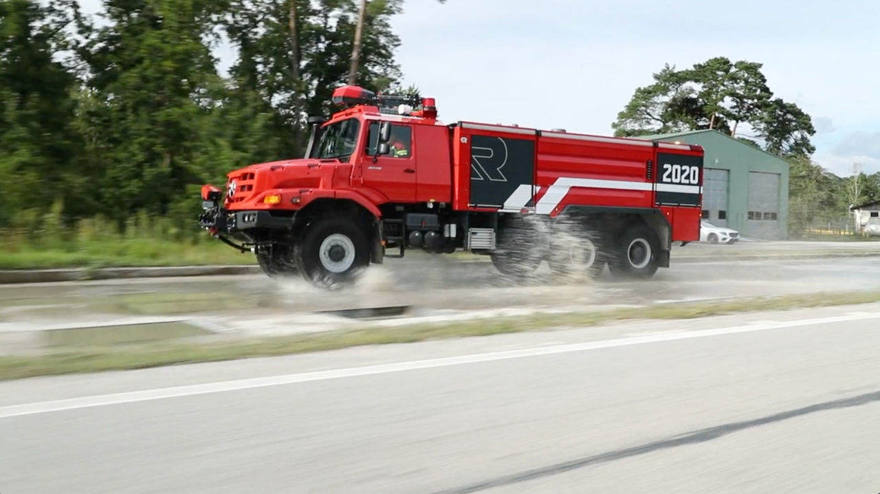 The image shows a red Unimog as an airport fire-fighting vehicle. The vehicle is rolling over a wet road. The body bears a large “R” and the year “2020”. A warehouse and trees can be seen in the background.