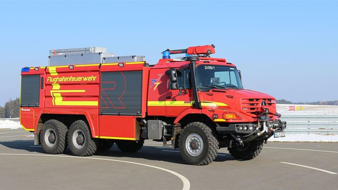 A large airport fire-fighting vehicle with “Flughafenfeuerwehr” (<i>Airport</i> <i>fire</i> <i>service</i>) on its side stands on an airport apron. The Zetros features several axles and large tyres, which suggests high off-road capability and a large extinguishing agent capacity. An asphalt road and snow is visible in the background.
