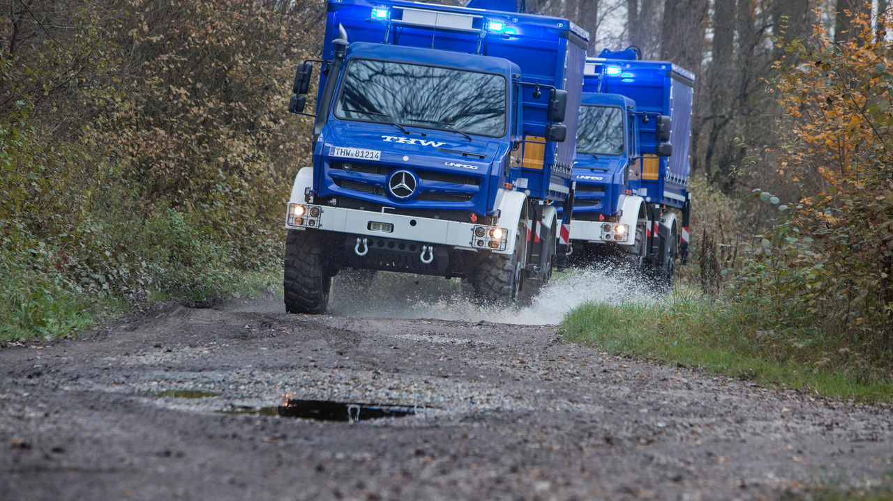 Two blue Unimogs with "TWH" written on them drive with their blue lights on along a muddy forest track. They are splashing up water.