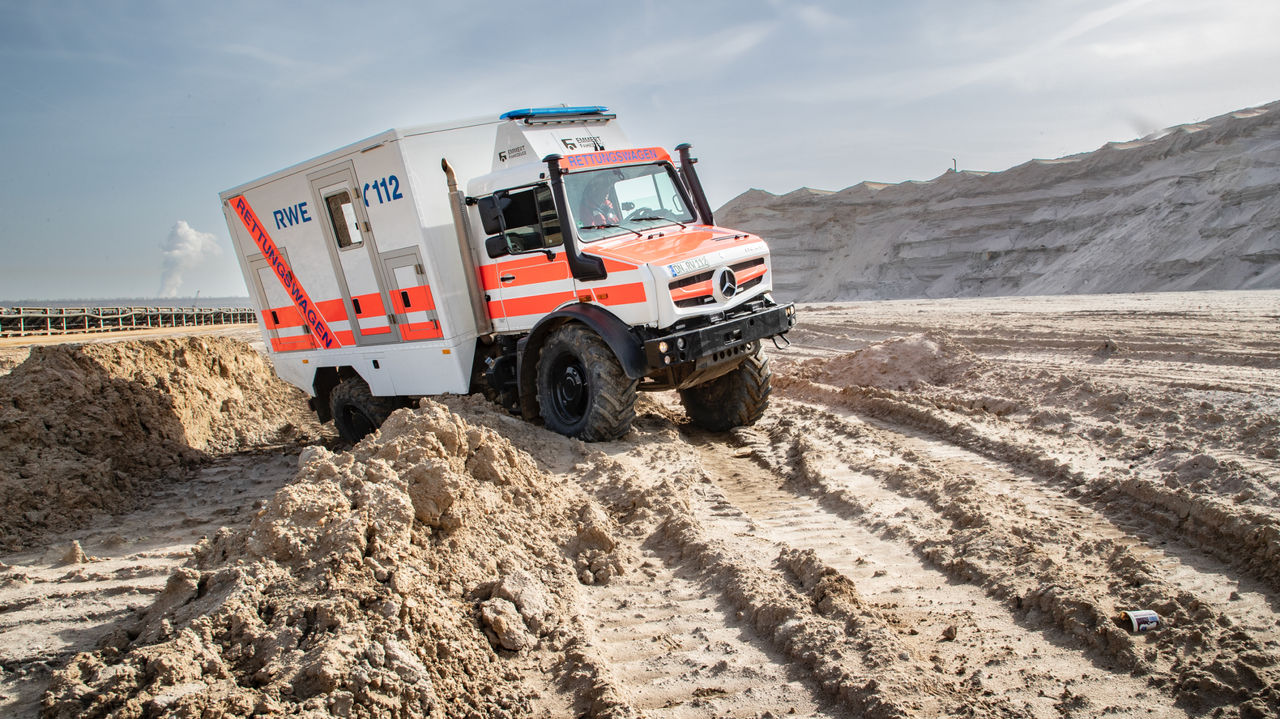 A white-and-orange rescue vehicle with "RWE" and "112" written on it makes its way across a sandy industrial site. The Unimog has large tyres and high ground clearance.