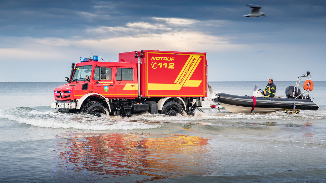 A red Unimog fire-fighting vehicle tows a rubber dinghy through shallow waters on a beach. A firefighter is steering the boat, while the vehicle has its blue lights on and has "NOTRUF 112" written on it. The sky is blue and there is an out-of-focus seagull in the foreground.