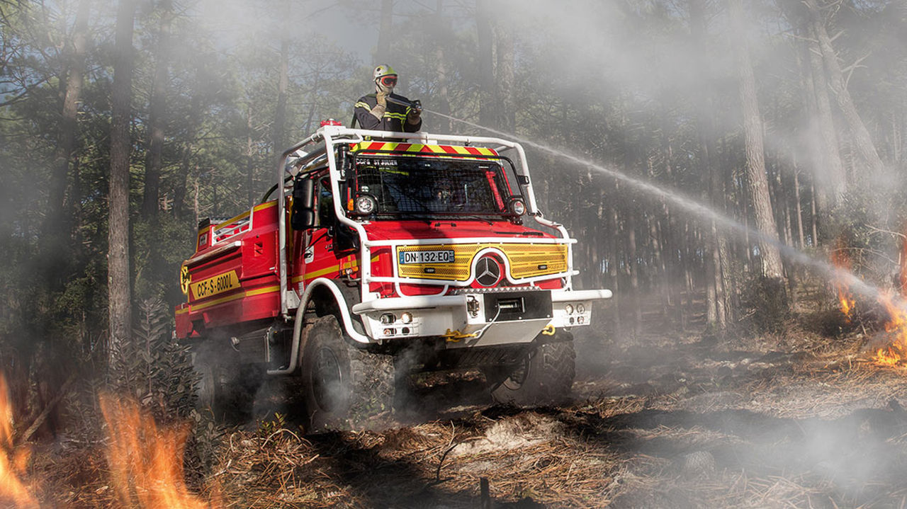 A red Unimog puts out a forest fire using its roof cannon. The forest fire-fighting vehicle is in a forest fire and a firefighter in protective clothing is operating the water cannon through the roof hatch. There is a protective cage in front of the windscreen.