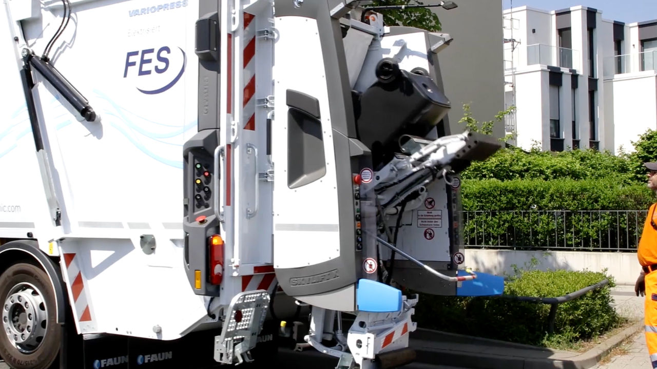 The image shows the rear of an Econic. This is a refuse collection vehicle with “FES” written on it. The truck’s lifting mechanism is extended and in motion. An employee in orange work clothes stands next to the rear of the vehicle. In the background is a modern residential building.