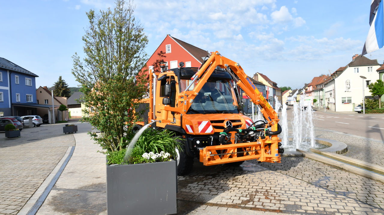 An orange Unimog with a crane arm mounted at the front of the truck waters a planter on a cobbled square. The Unimog has a snowplough linkage at the front, but it is not in use. In the background is a fountain, houses and parked cars. 