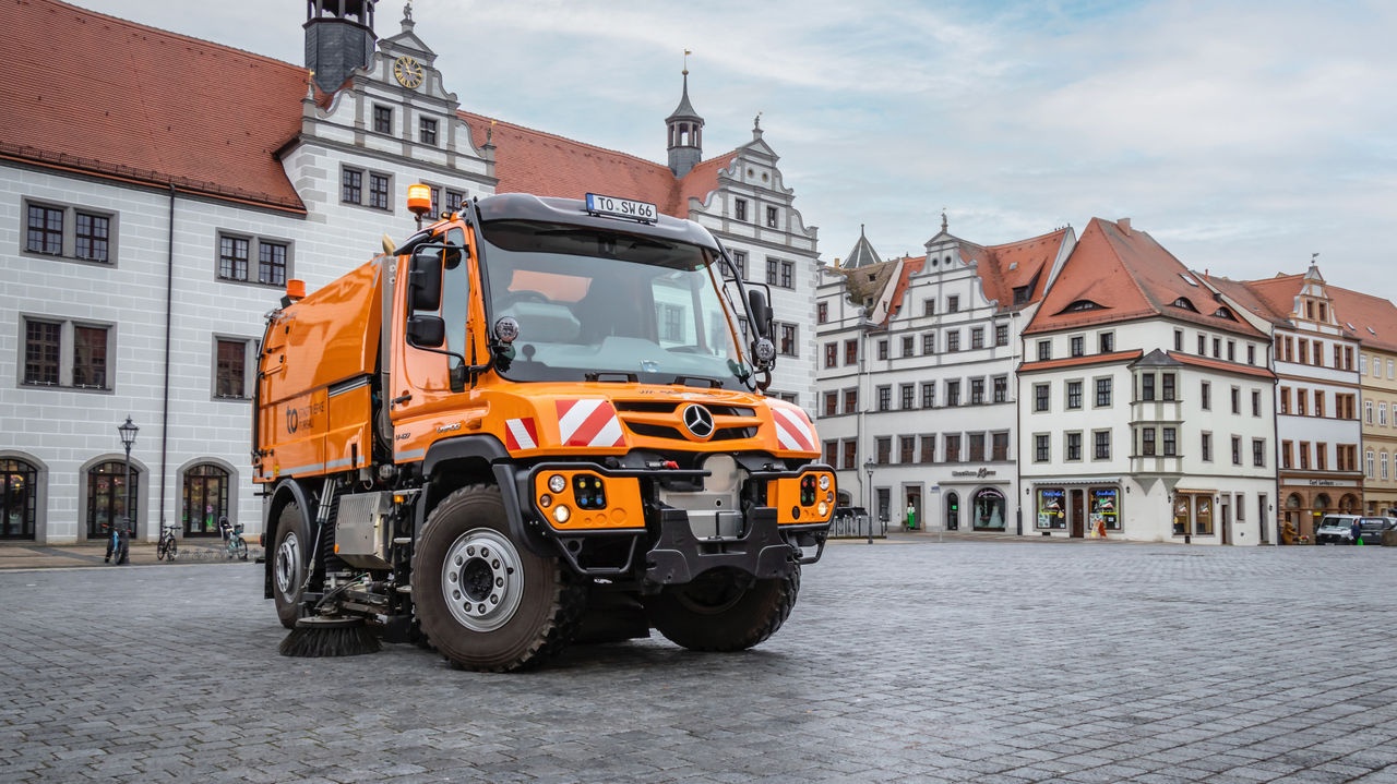 An orange Unimog with a road sweeper is parked in a cobbled square. In the background are historic buildings with white façades and red roofs. The square seems empty, with just the Unimog standing there. 
