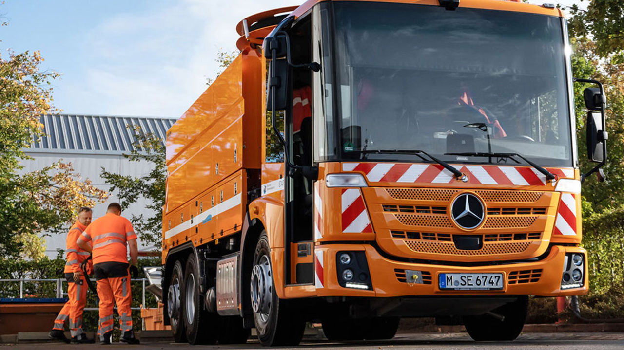 An orange Econic refuse truck is parked on a road. Two workers in orange protective clothing stand behind a container at the rear of the vehicle. In the background are trees and a building with a grey corrugated sheet metal roof.