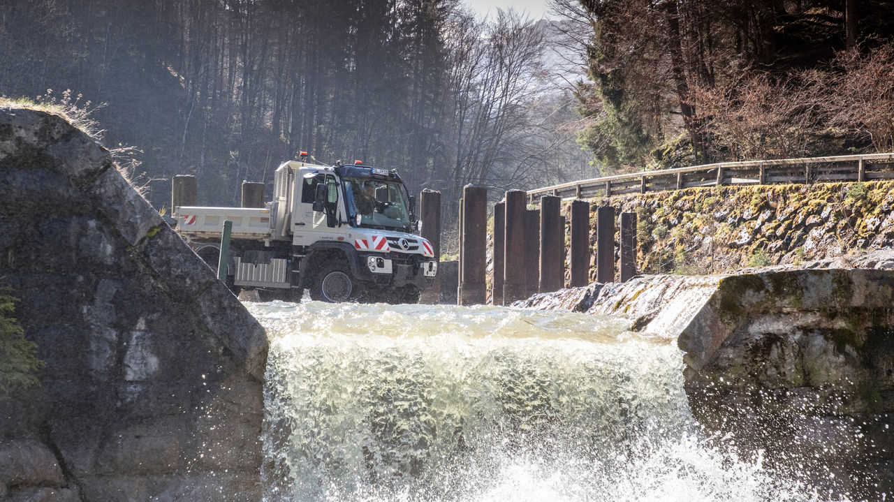 A Unimog crosses a small weir or waterfall. The water splashes around the wheels of the vehicle. The Unimog has a white cab and a grey loading surface. In the background is a wooded landscape. A stone wall and wooden posts frame the weir.