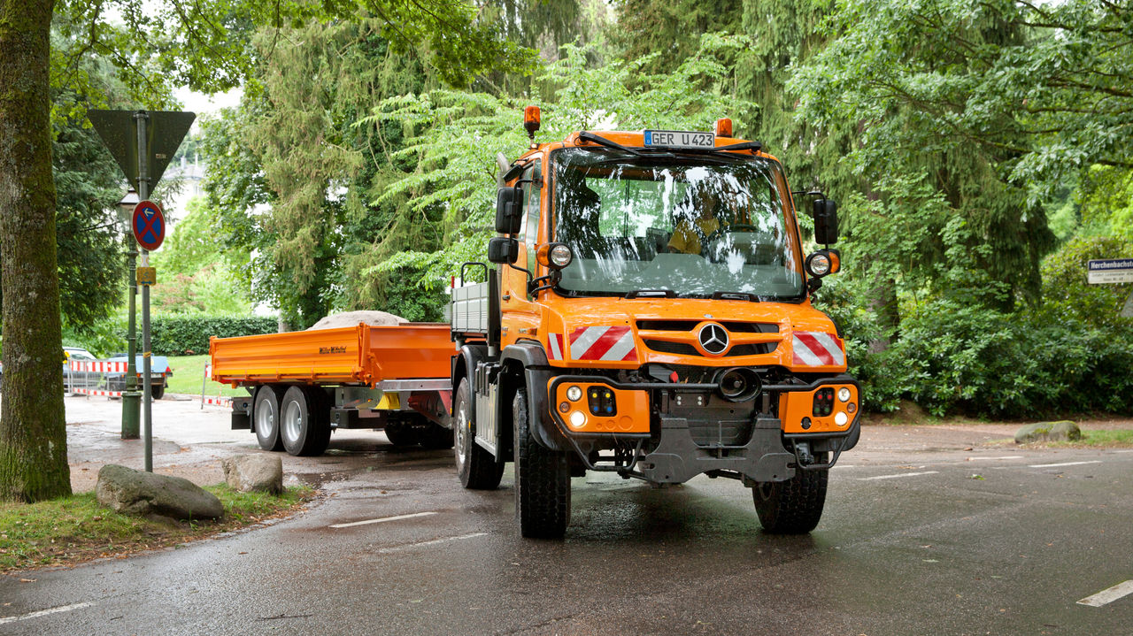 An orange Unimog pulls an orange flatbed trailer along an asphalt road. The truck is at a junction, surrounded by trees and shrubs. Other parked vehicles and traffic signs are visible in the background. The road surface seems to be damp.
