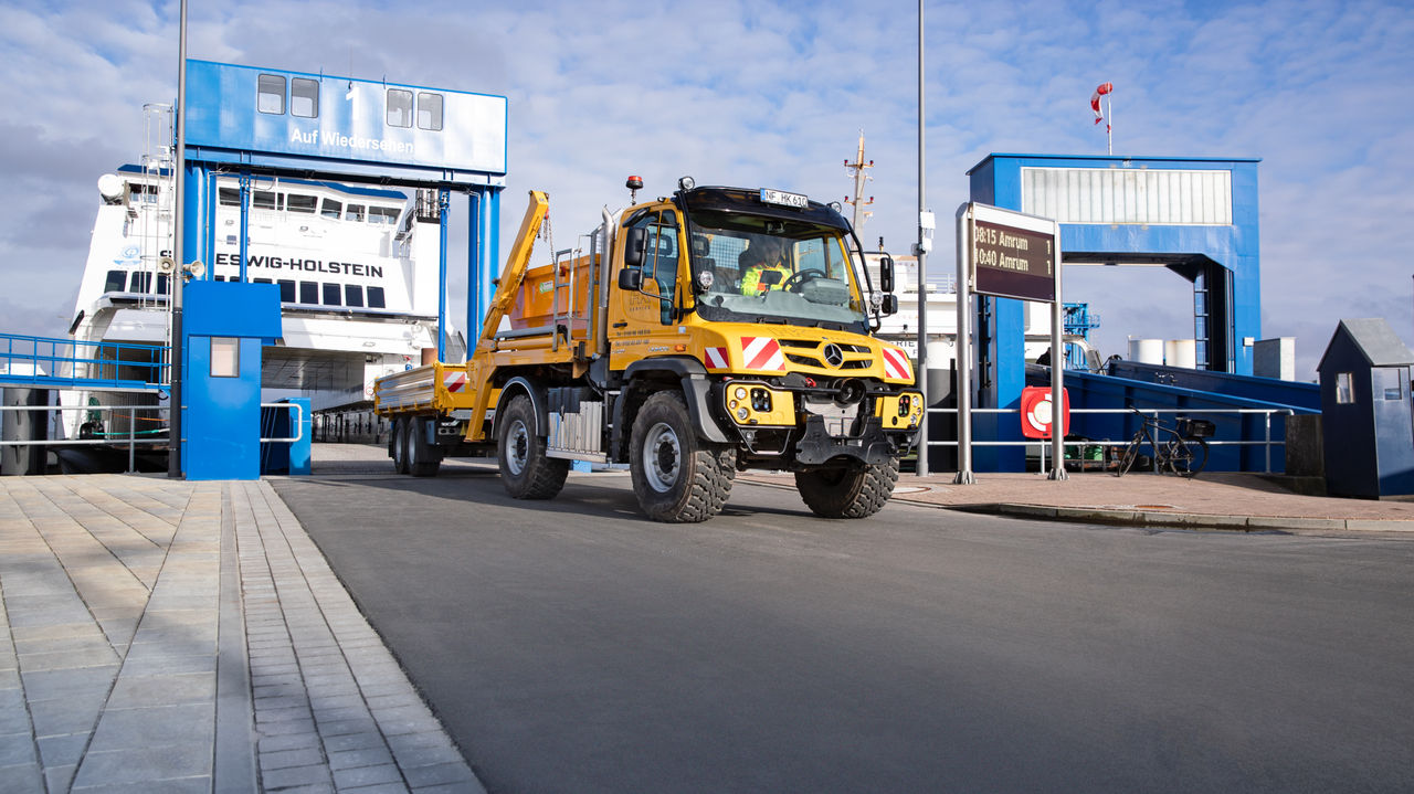 A yellow Unimog with a trailer drives off a ferry. The ferry, with “SCHLESWIG-HOLSTEIN” written on it, is moored at a blue ferry dock. The area in front of the dock is asphalted, and there is a paved walkway next to it.