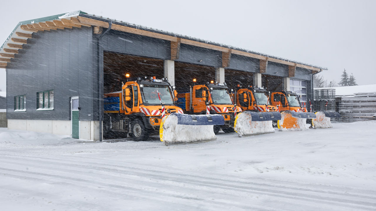 Three orange snowploughs from Mercedes-Benz are ready to drive away in a vehicle hall. The snowploughs are equipped with dozer blades and the surrounding area is covered in snow. It is snowing heavily.